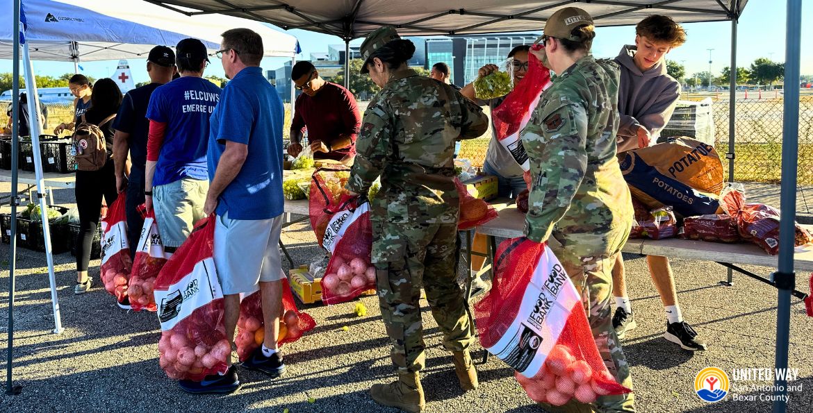 Adults stand in a line holding mesh bags full of vegetables next to tents and tables.