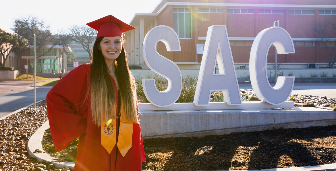a woman in a red graduation cap and gown smiles in front of the San Antonio College 